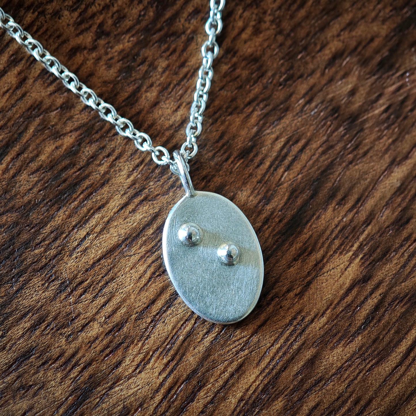 Blindenschrift-Kettenanhänger oval mit einzelnem Buchstaben, Nahaufnahme auf dunklem Holz