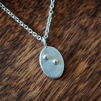 Blindenschrift-Kettenanhänger oval mit einzelnem Buchstaben, Nahaufnahme auf dunklem Holz