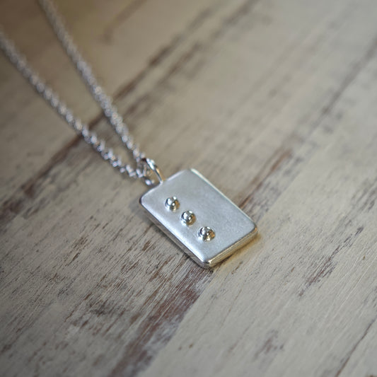Blindenschrift-Schmuck, Nahaufnahme von Kettenanhänger mit Buchstabe in erhabener Punktschrift, lesbare Braille-Buchstaben als Kette